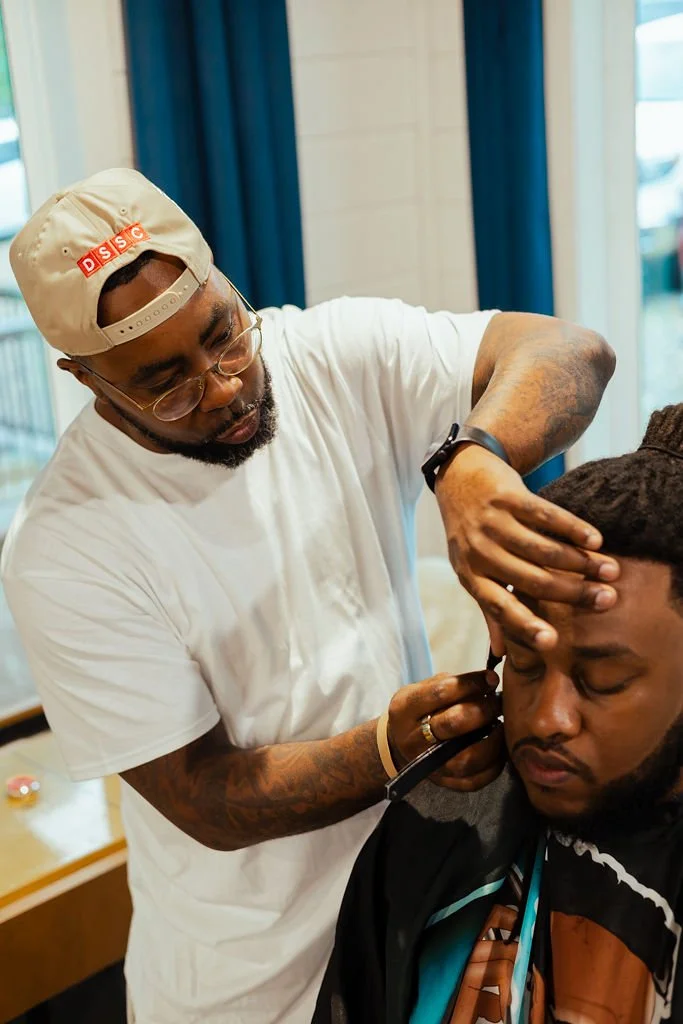 A barber wearing glasses, a beige cap, and a white t-shirt is giving a haircut to a man with dreadlocks, who has his eyes closed, in a salon with blue curtains in the background.
