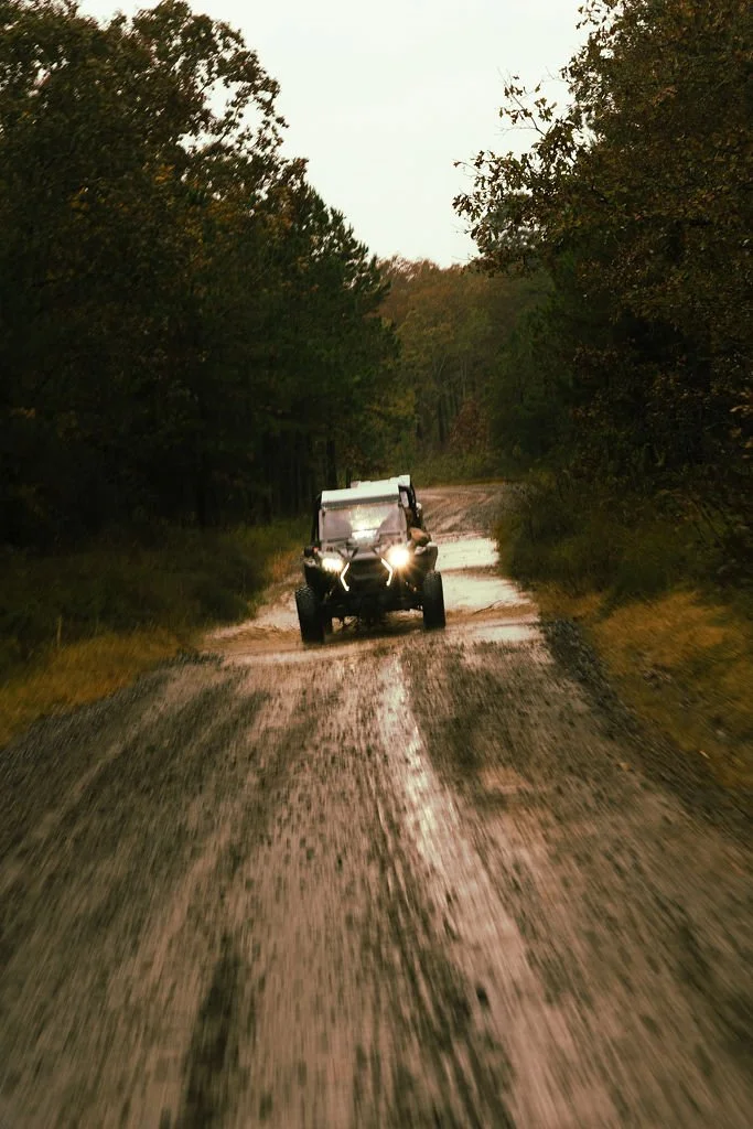 An off-road vehicle driving on a dirt road surrounded by trees in a forest during dusk or early evening.
