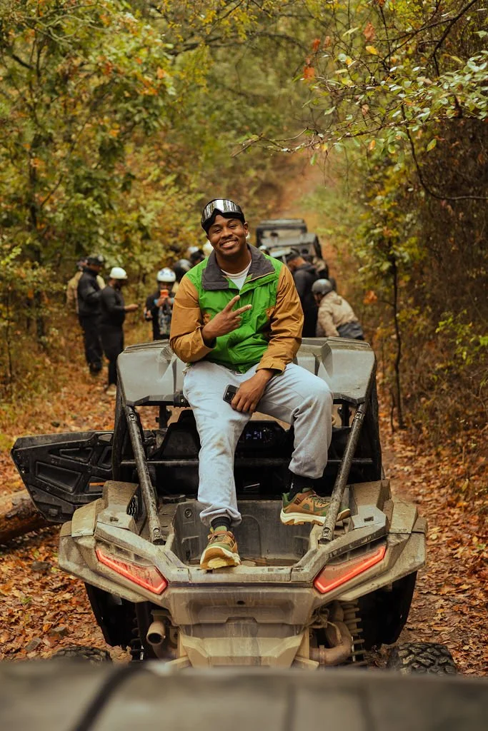 A young man smiling and making a peace sign while sitting on a rugged all-terrain vehicle in a forested area during fall, with trees and other people on ATVs in the background.