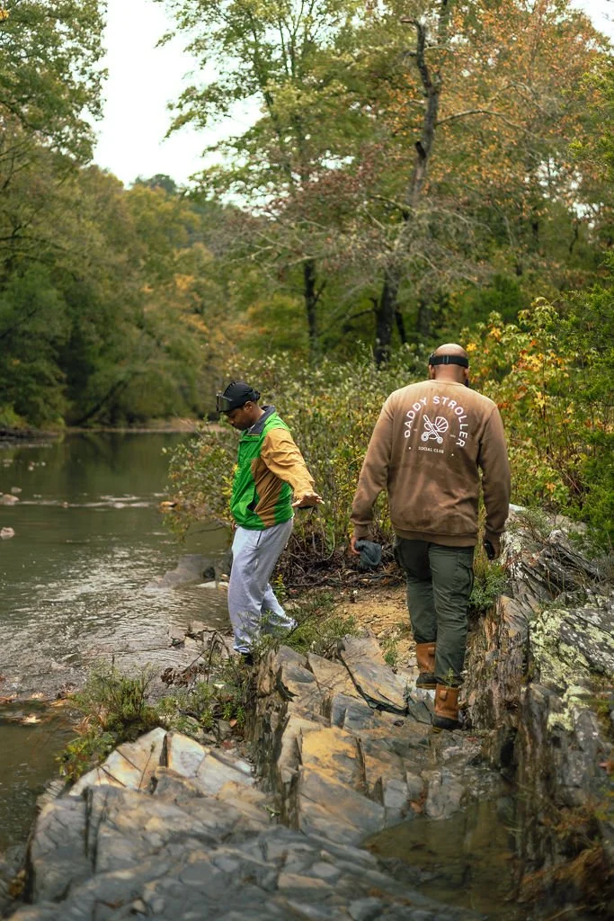 Two people standing on rocks by a river in a forest, one wearing a green and brown jacket and the other in a tan sweatshirt with 'Daddy Stroller Social Club' logo, both with headwear, surrounded by green and autumn-colored trees.
