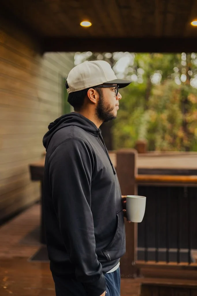 Side profile of a man wearing a black hoodie, glasses, and a gray cap, holding a white mug on a wooden balcony or porch with trees in the background.