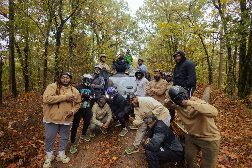 Group of people in casual and motorcycle gear gathered in a wooded area with autumn leaves on the ground, some sitting on the ground, others standing or sitting on a vehicle.