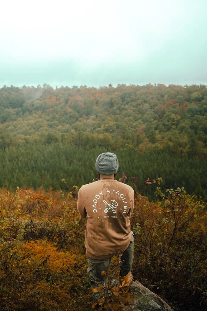 Person wearing a gray beanie and tan jacket with "Daddy Stroller Social Club" logo, standing on a rock and looking at a forested hillside with fall foliage.