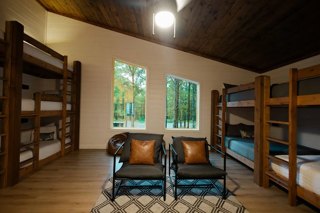 A cozy bedroom with two sets of wooden bunk beds, two black chairs with brown pillows, a small table, and two large windows overlooking a wooded outdoor scene.