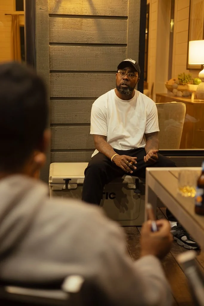 A man with a beard wearing glasses, a white t-shirt, and a black cap is sitting on a cooler, speaking to two people in the foreground, one of whom is holding a phone.