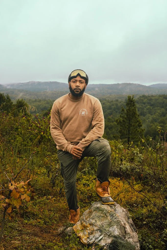A man with a beard wearing a tan sweatshirt, gray pants, and brown boots standing on a large rock in a forested area with rolling hills in the background.