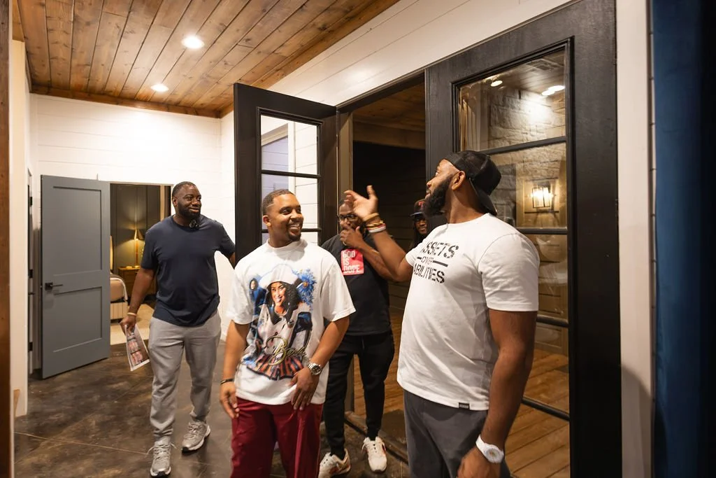 Group of five men laughing and talking inside a modern house, some wearing casual clothing, one with a graphic t-shirt, in front of black-framed glass doors and a wooden ceiling.