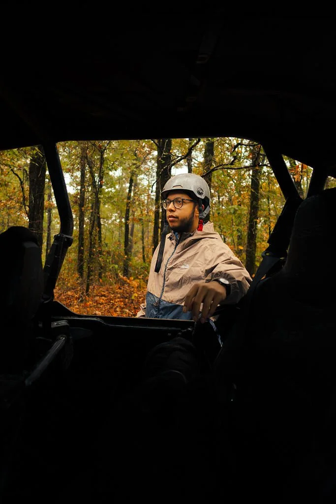 A man wearing a helmet and glasses stands outside of a vehicle in a wooded area with autumn leaves.