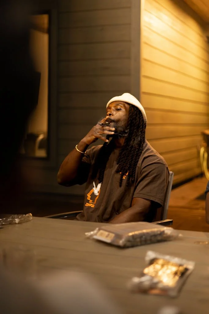 A man with dreadlocks wearing a white hat and casual dark t-shirt is sitting at a table, smoking a cigarette, in a warmly lit indoor or outdoor setting with wooden walls.