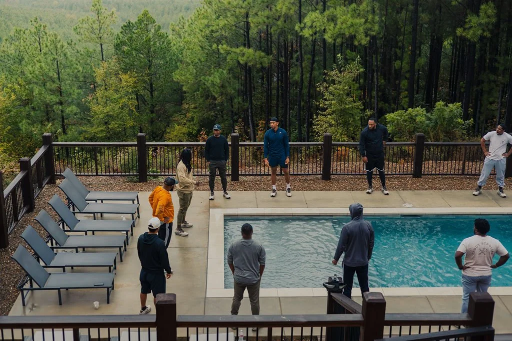 A group of people standing around a pool on a deck, with a wooded forest background.