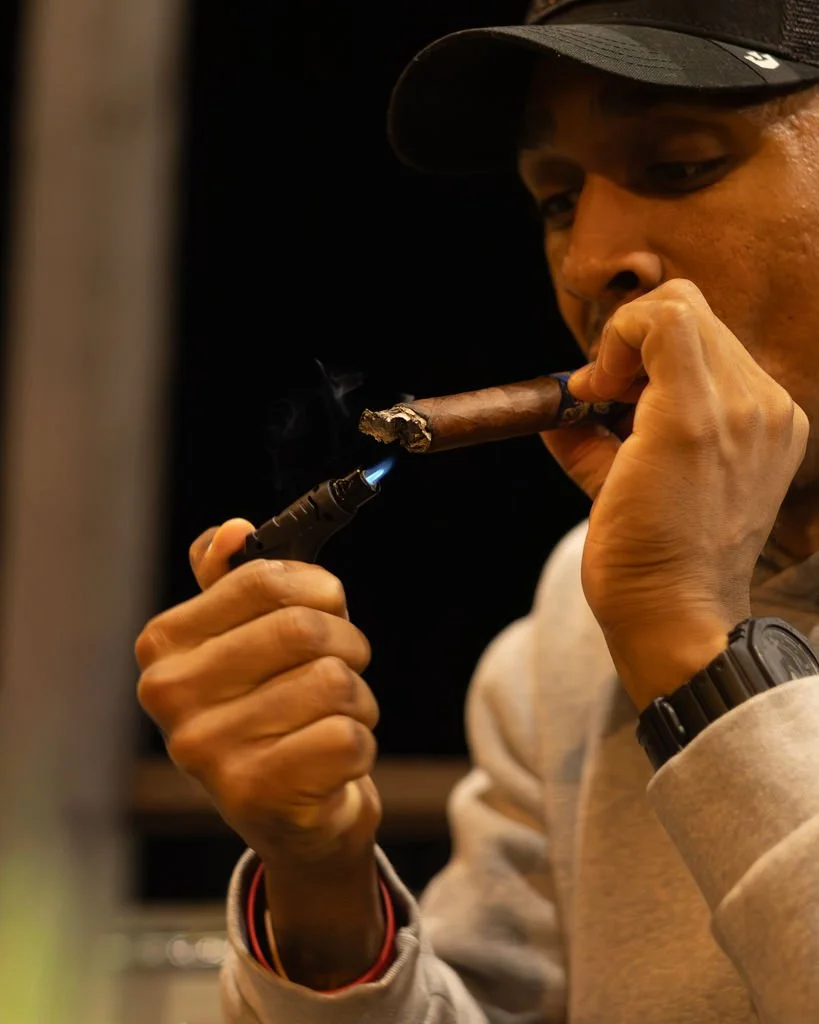A man wearing a black cap holds a lit cigar in his mouth while using a small blowtorch to light it.