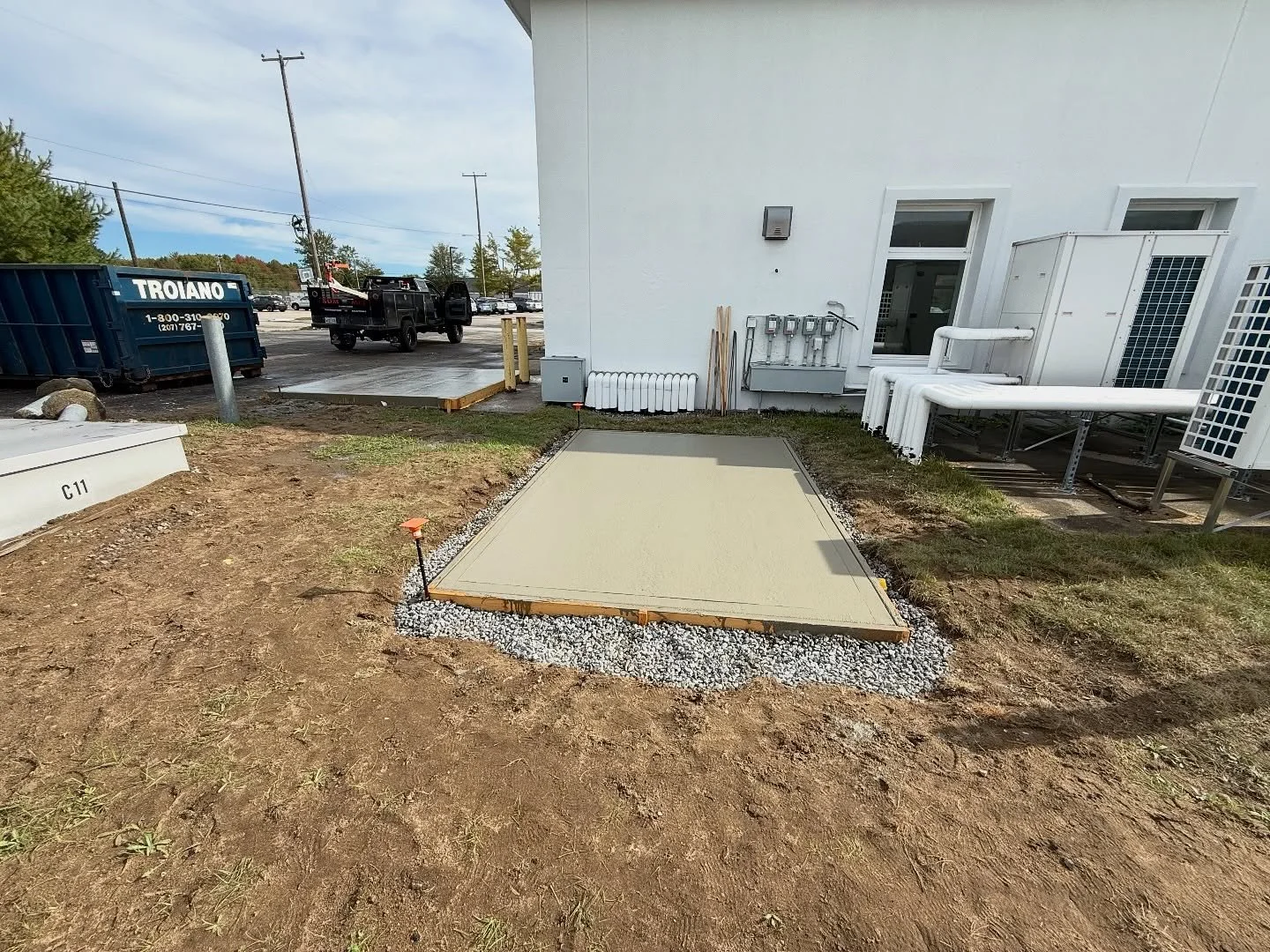Concrete slab freshly poured at construction site, surrounded by gravel, with surrounding dirt and construction equipment in background.