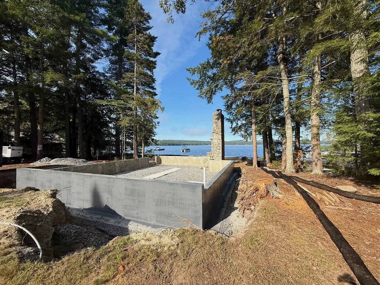 Construction site near a lake with a partially built concrete structure made of cinder blocks, tall trees surrounding the area, and boats visible on the water in the background.
