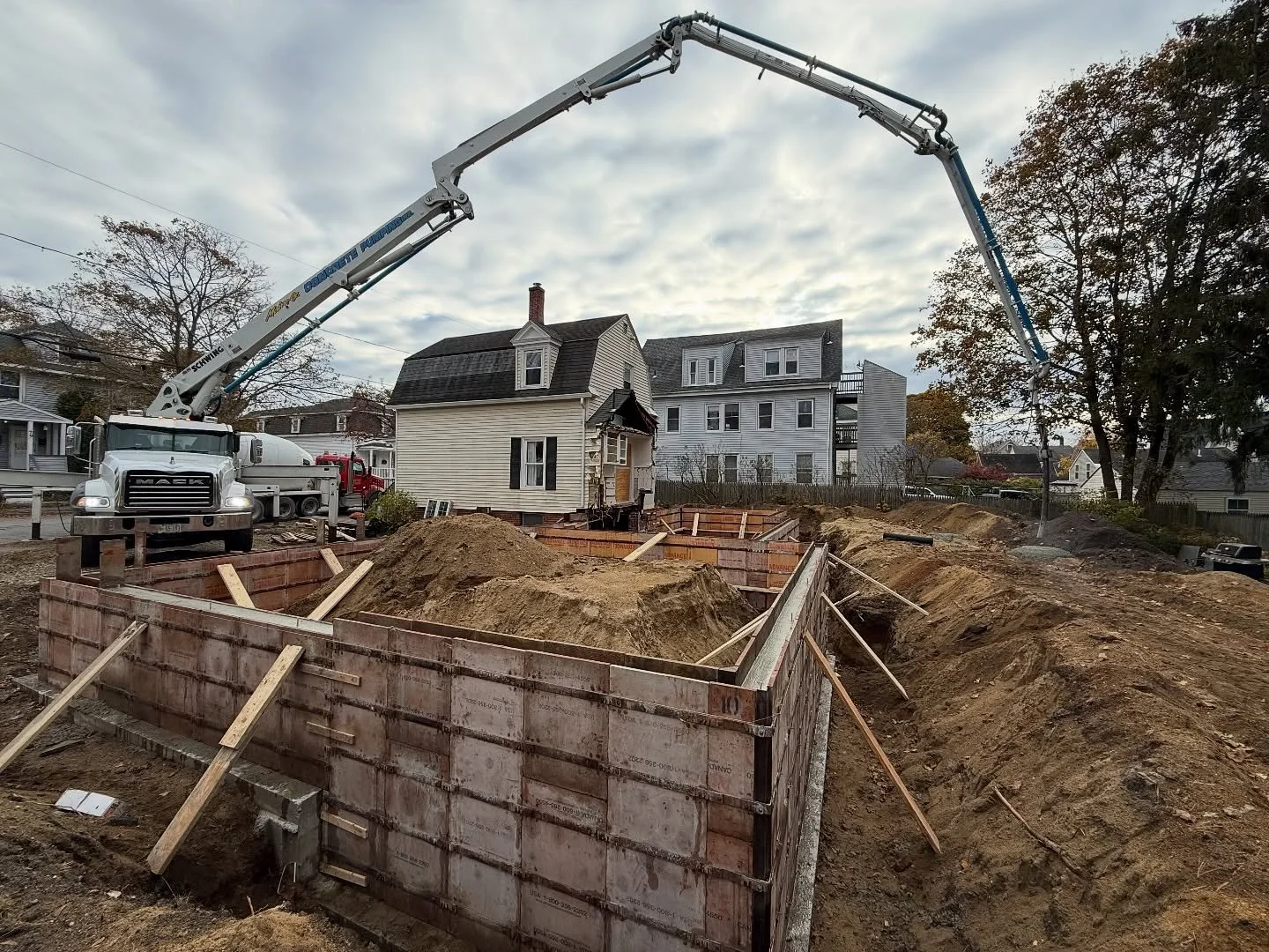 Construction site with foundation walls in progress behind a large concrete pump truck parked on the street, with residential houses and trees in the background, under a cloudy sky.