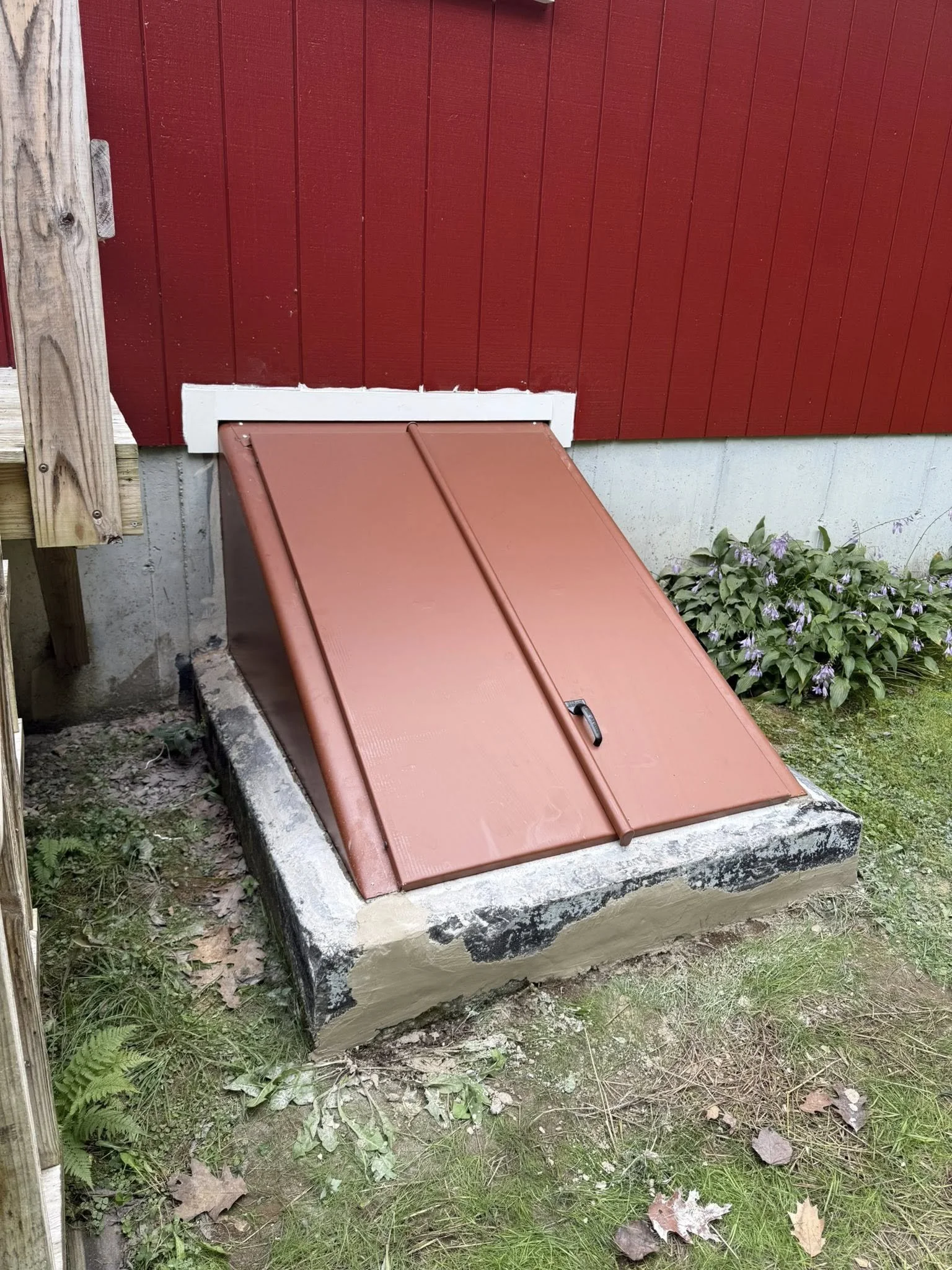 A red outdoor hatch with a handle, installed on a concrete foundation next to a red wooden wall, with purple flowers and green plants nearby.