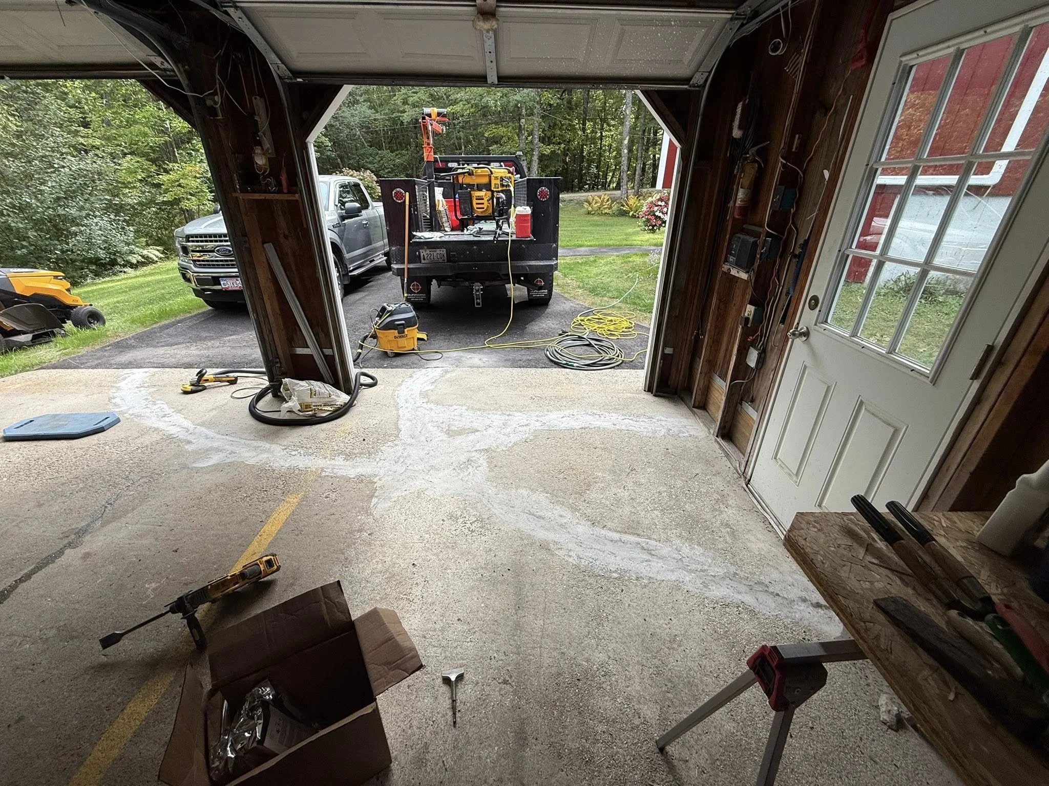 View of a garage with the door open showing a driveway outside with work vehicles, tools, and equipment for a project in progress.