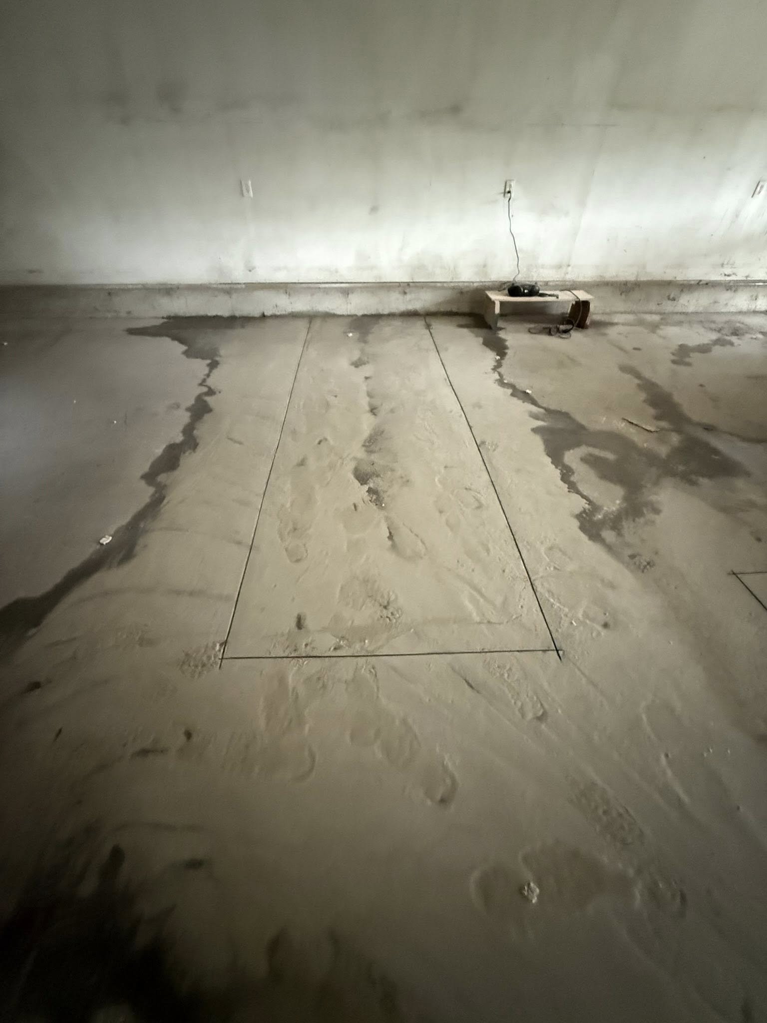 Bare concrete floor with chalk markings and wet patches, against a wall with an electrical outlet and a wooden shelf holding a black device and cords.