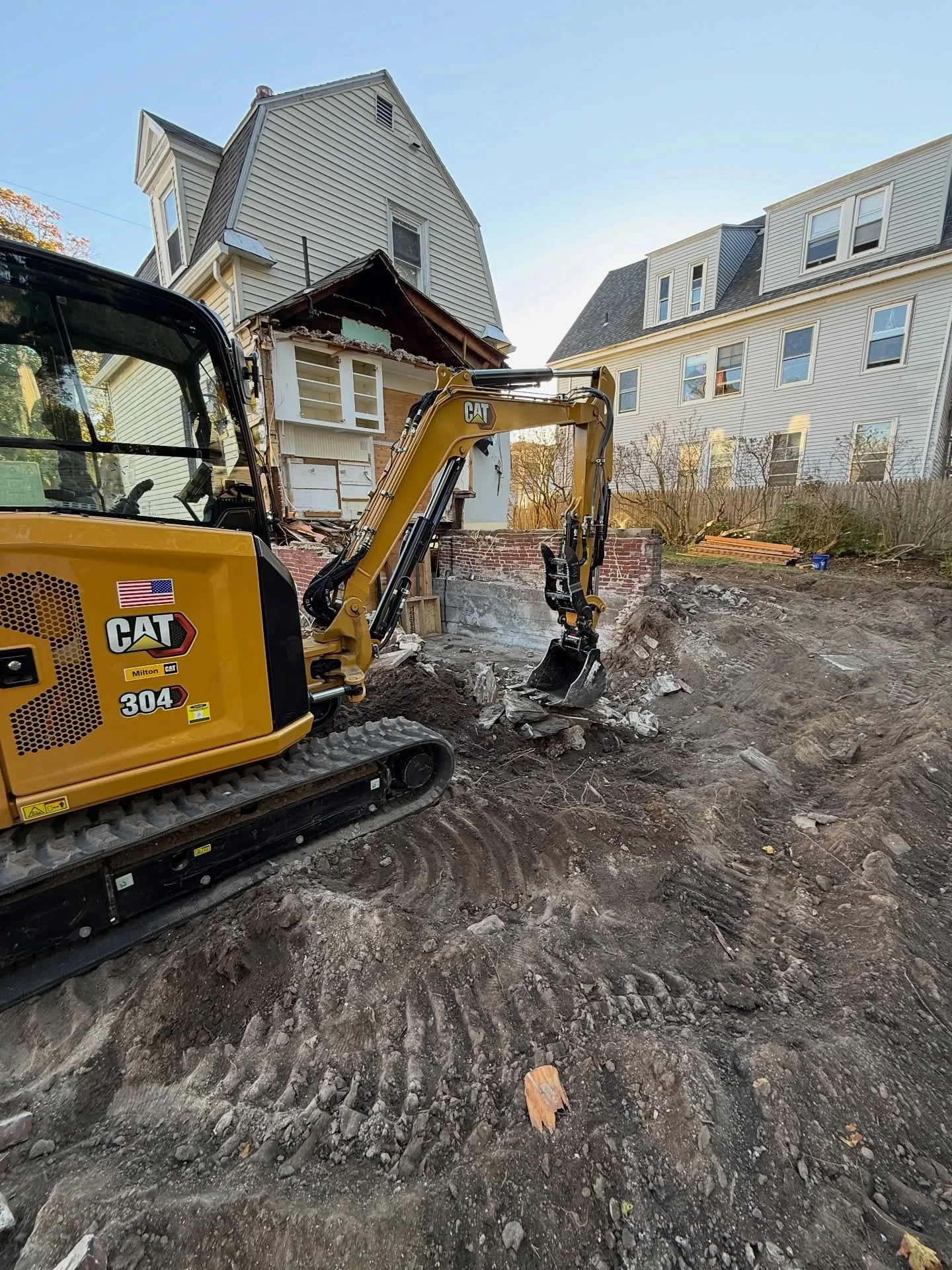 A small yellow Caterpillar excavator working on a house demolition or construction site, with neighboring houses visible in the background.