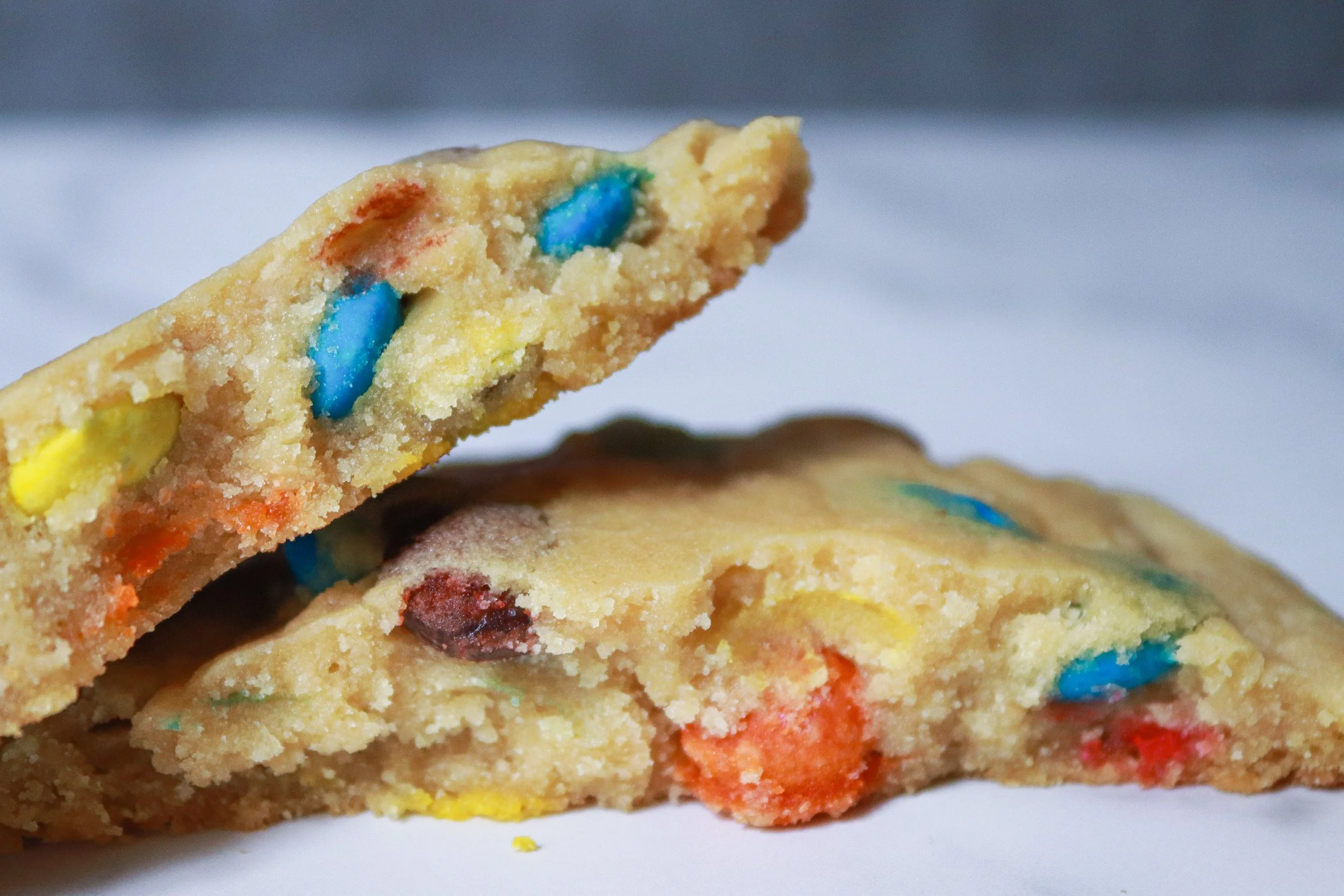 Close-up of two colorful M&M cookies with rainbow-colored candy pieces on a white surface.