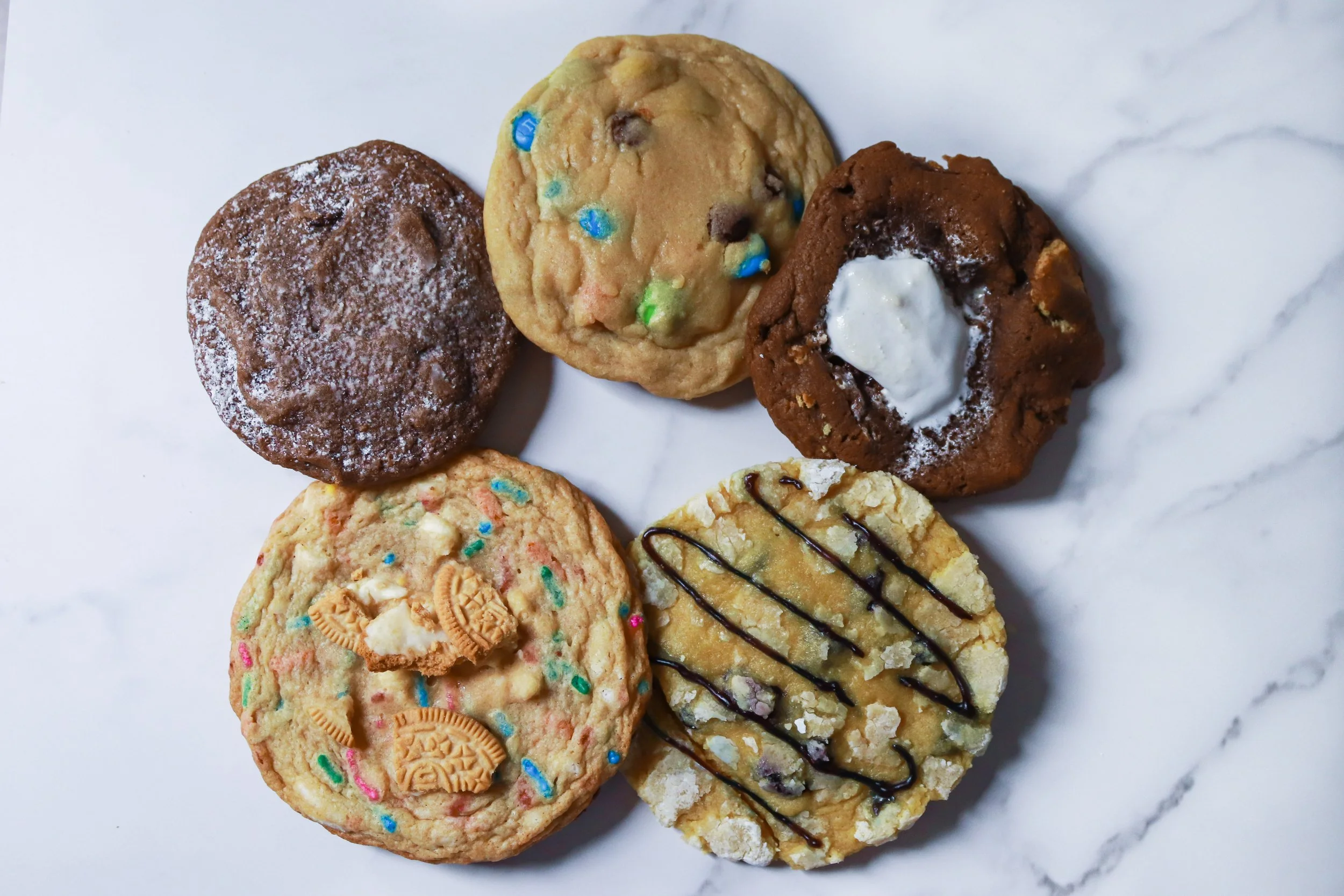 Five assorted cookies on a white marble surface: a chocolate cookie with chocolate filling, a cookie with colorful sprinkles and Oreo pieces, a S'mores cookie, an M&M cookie, and a lemon cookie with a blueberry drizzle.