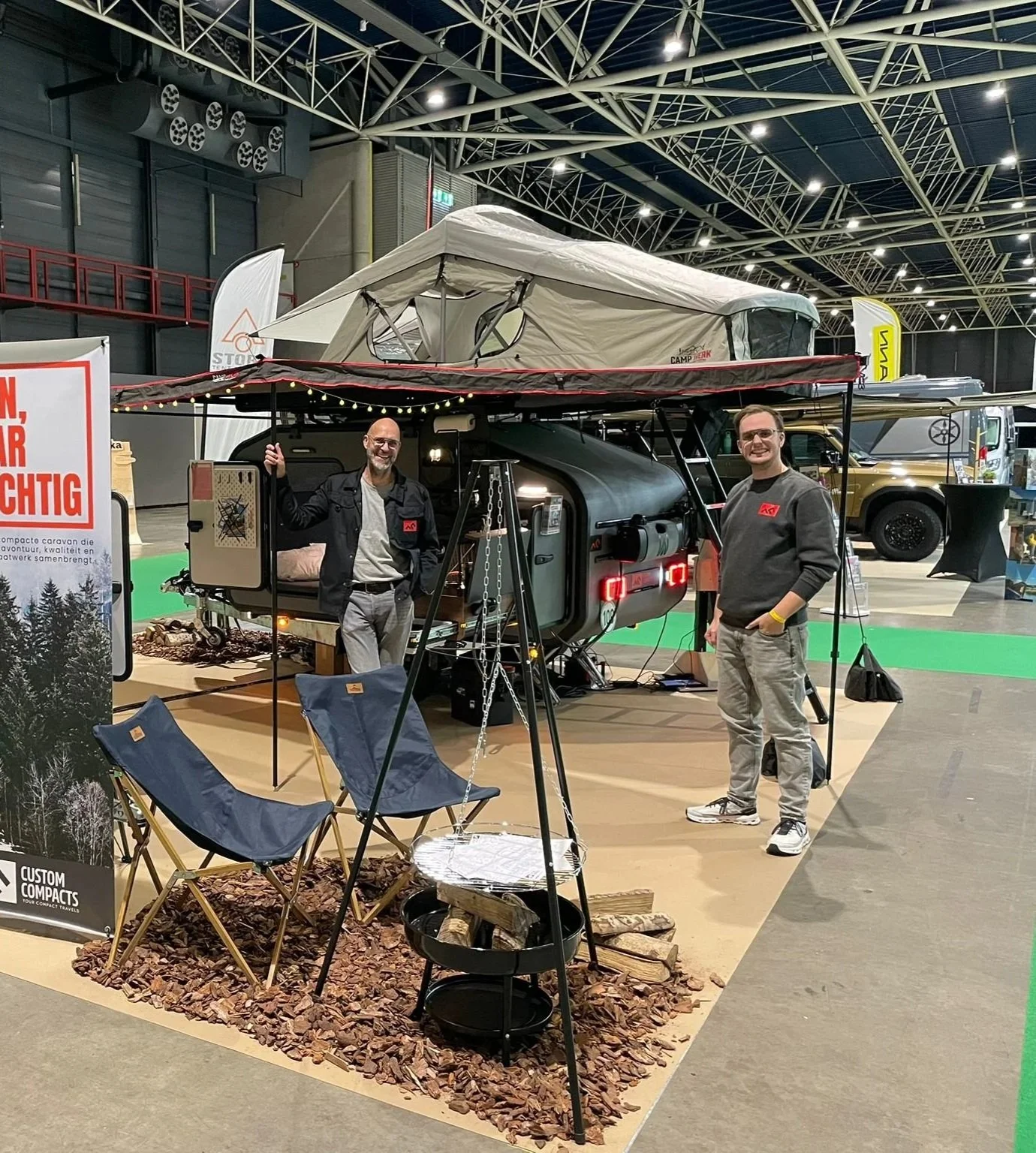 Twee mannen staan naast een opblaasbare tent op een caravan, beide glimlachend, in een binnenhal met andere caravan en uitstallingen.