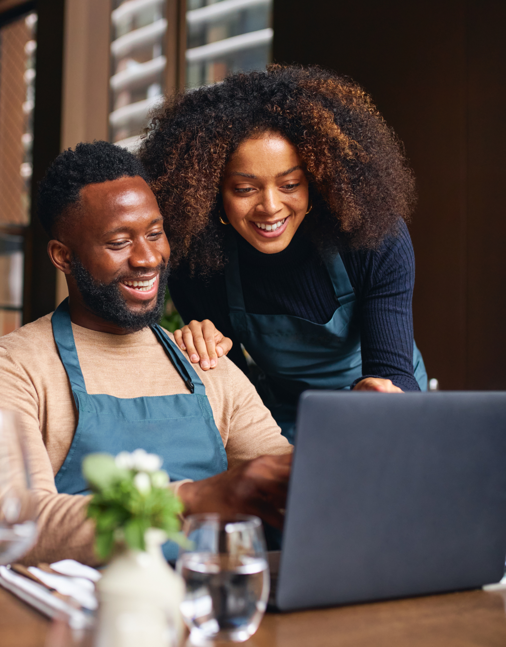 A smiling man and woman wearing aprons looking at a laptop screen, in a restaurant or cafe setting.