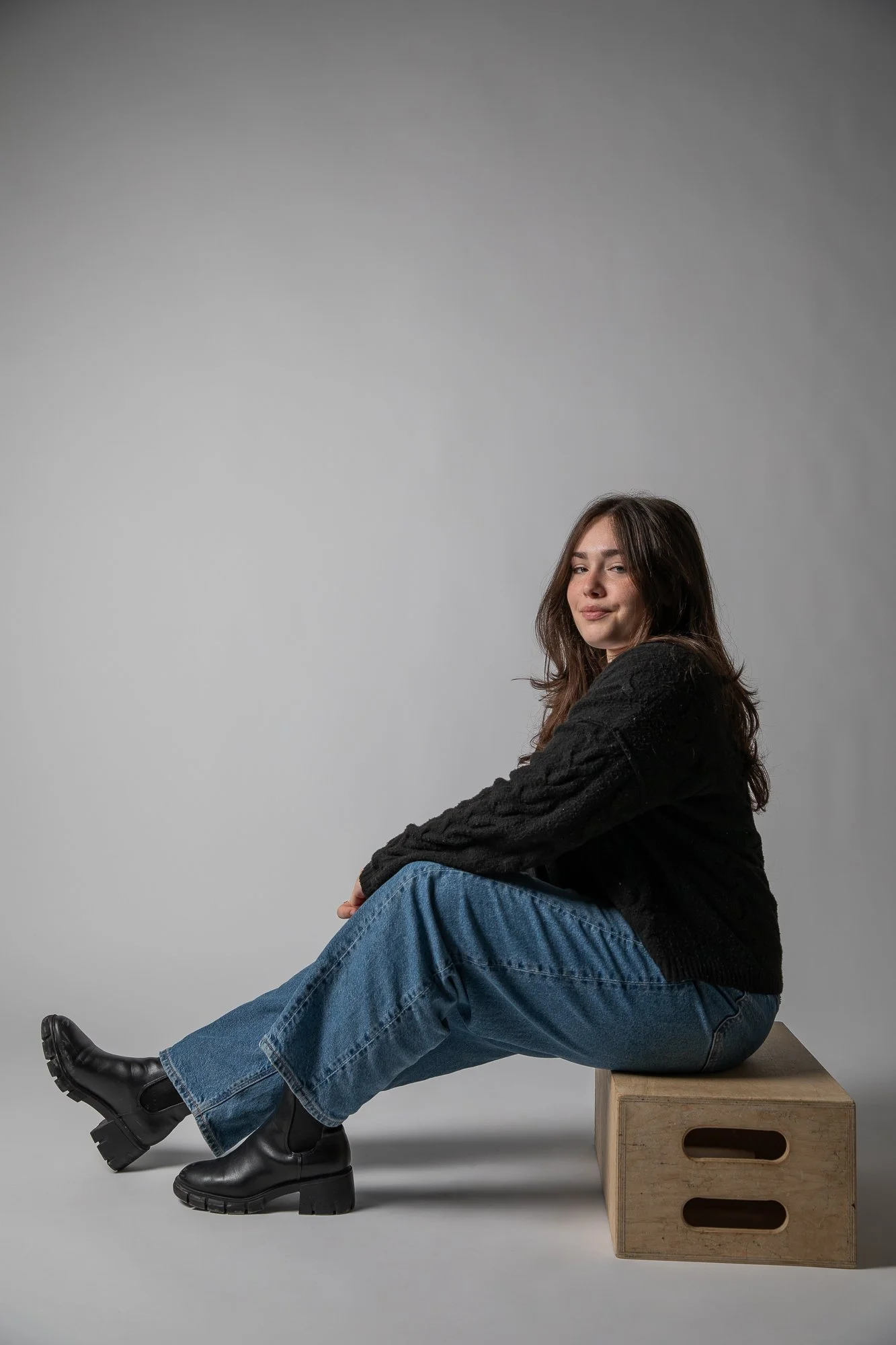 A person with long hair wearing a black sweater, blue jeans, and black boots, sitting on a wooden box against a gray background.