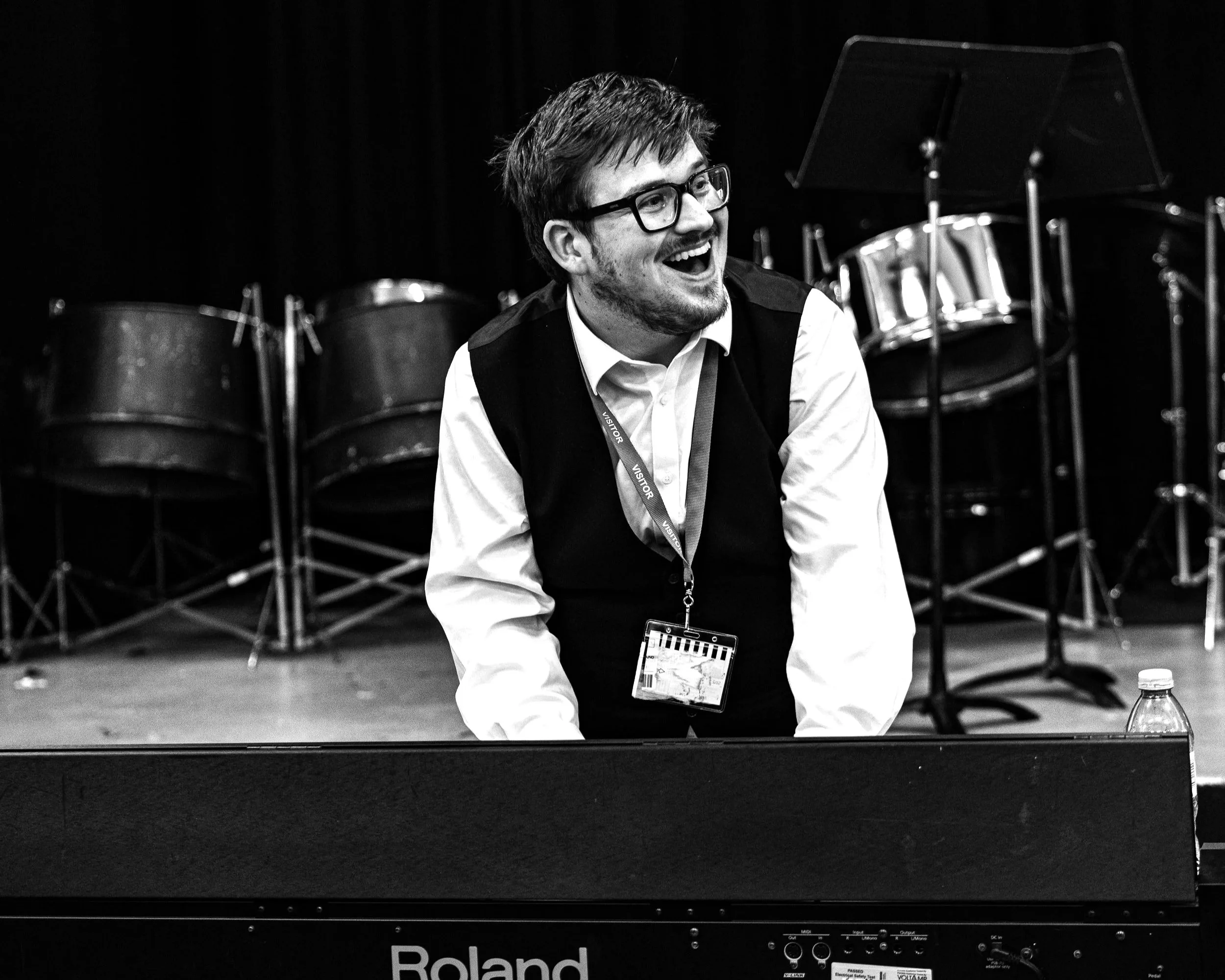 A man with glasses, a beard, and a big smile playing a Roland keyboard during a musical event, with drums and music stands in the background.