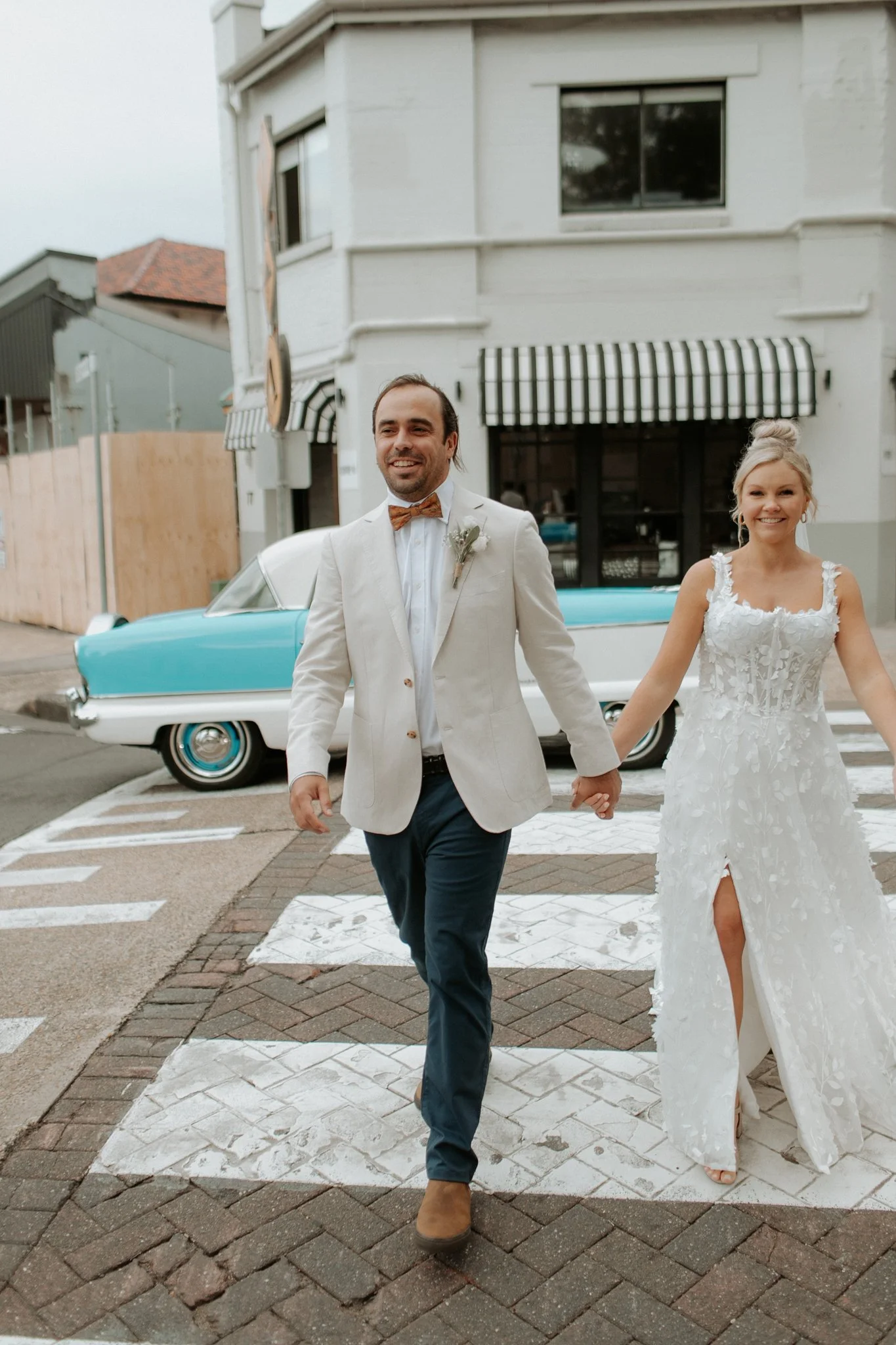 A couple in wedding attire walking hand in hand on a crosswalk, with a vintage car and buildings in the background.