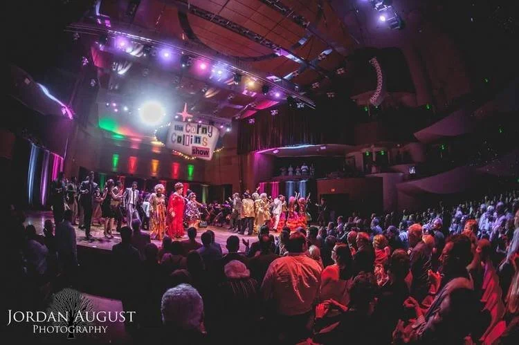 A lively stage performance with a large audience in a theater. Costumed performers are gathered under spotlights, showcasing a vibrant scene. A sign reads 'The Corny Collins Show' indicating a theatrical production. Colorful lighting adds drama to th