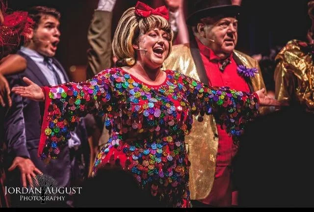Performers on stage in colorful costumes, with one in a sequined dress and big bow, singing enthusiastically. Photography credit to Jordan August.