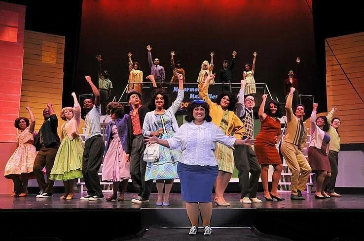 A theatrical stage performance with a diverse cast of performers in 1960s-style clothing. The actors, positioned in various poses, fill the stage with lively expressions and gestures, suggesting a musical number. The background features steps and sim