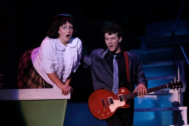 A stage performance featuring a woman leaning over a ledge, appearing surprised, and a man standing next to her holding a guitar, smiling. The background is dark, with stage lighting illuminating the performers.