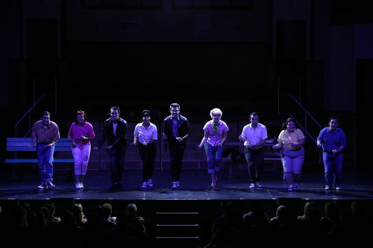 Group of performers on a stage under dim lighting, dressed in various costumes, in the midst of a theatrical performance from Grease the musical.