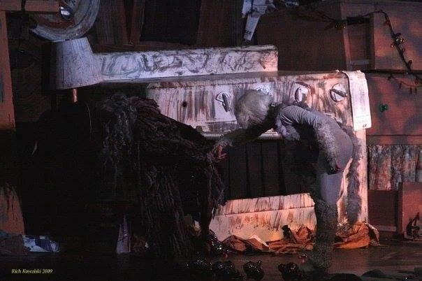 A person in theatrical costume interacting with stage props, including a stove and other kitchen items, in a dimly lit setting.