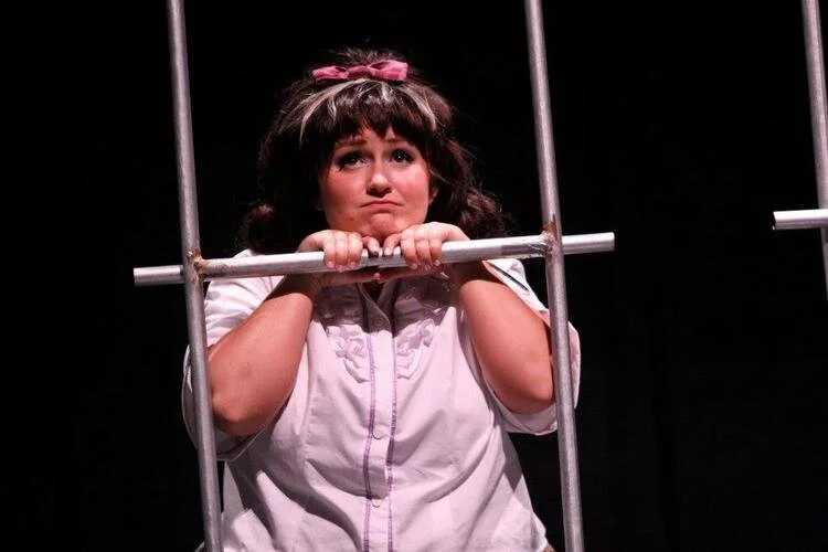 Person dressed in 1960s style, with a pink bow, peering through metal bars, performing as Tracy Turnblad in a theatrical production of Hairspray.
