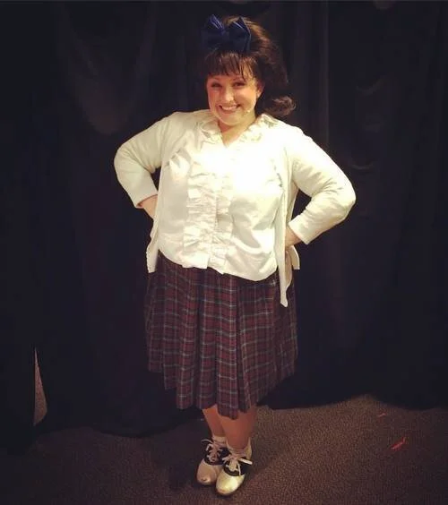Person in a white shirt and plaid skirt posing in front of a dark background