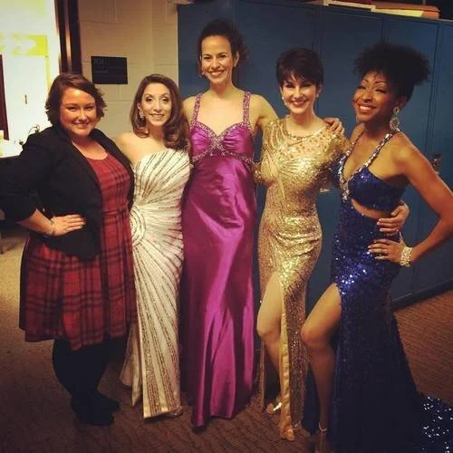 Five women posing in glamorous evening gowns; four are wearing formal dresses in various colors including white, purple, gold, and blue, while one is wearing a red plaid dress with a black jacket. They are standing together indoors, in front of blue 
