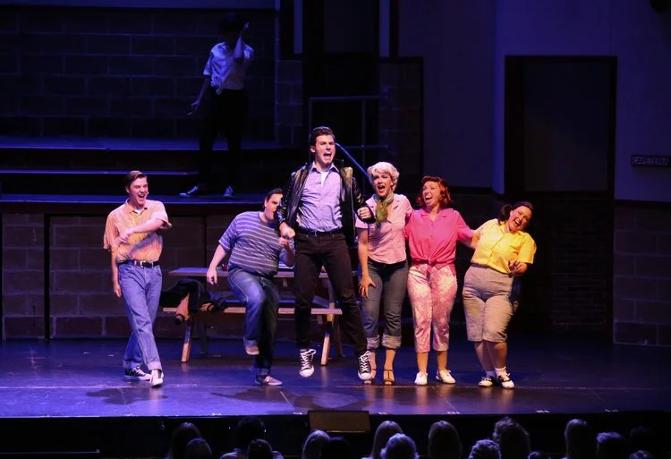 Group of actors on stage performing a musical number from Grease the musical.
