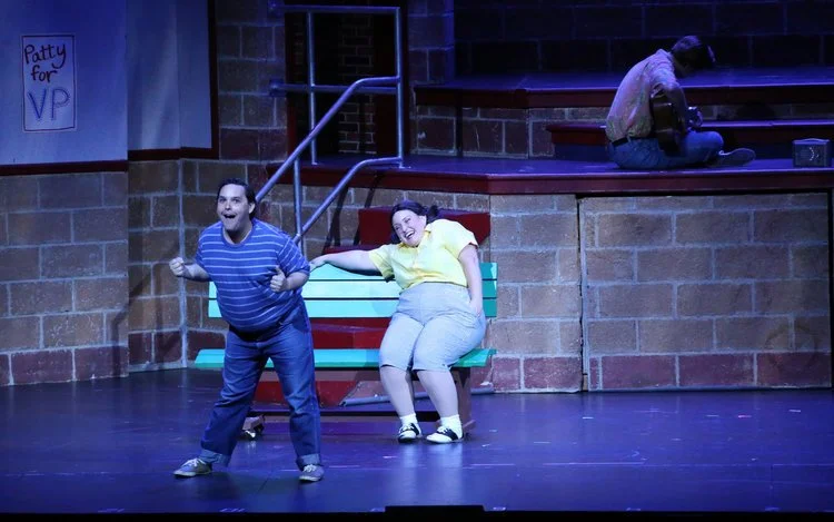 Two actors on stage in a theatrical performance, one standing and one sitting on a bench. The standing actor wears a blue striped shirt, and the seated actor in a yellow top looks amused. Background features brick walls and a sign saying "Patty for V
