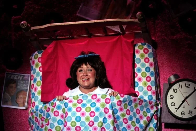 A woman with dark hair and a blue bow lies in bed with a colorful polka-dot blanket and a red headboard. A clock shows 10:10 beside her. Teen magazines and records are visible in the background.