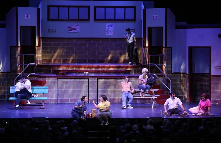 Stage play scene with actors seated on benches, depicting a school environment with brick walls, staircases, and exits, during a performance.