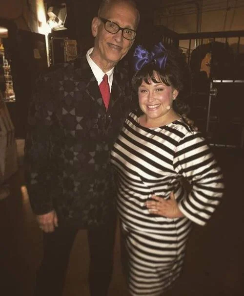 Two people standing together; one wearing a patterned suit and red tie, the other in a black and white striped dress with a purple bow in their hair.