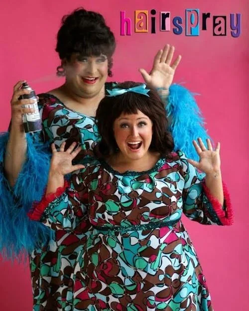 Two people dressed in colorful costumes with feathered accents, posing playfully in front of a pink backdrop, holding a can of hairspray with the word "Hairspray" written above.