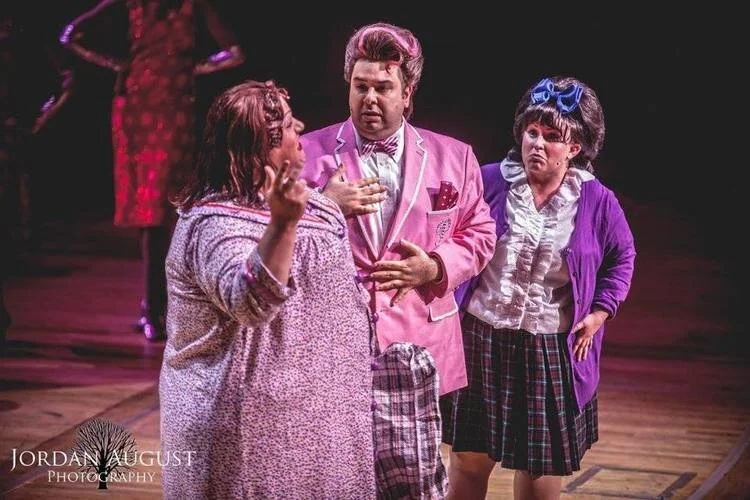 Three actors on stage in vivid costumes, performing a scene from a theatrical production of Hairspray.