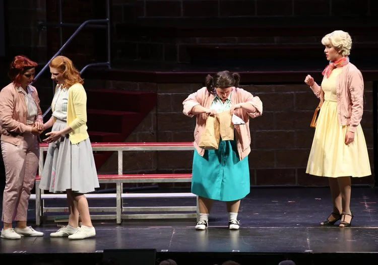 Four women on stage dressed in mid-20th century fashion, with 1950s-style hair and outfits, standing near bleachers. One holds a paper bag.