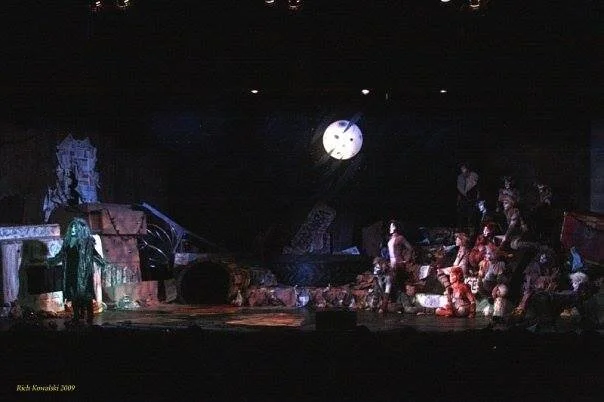 A theater stage set resembling an urban alley, featuring actors in colorful costumes, with a large clock in the background. The scene is dimly lit, creating a mysterious atmosphere.