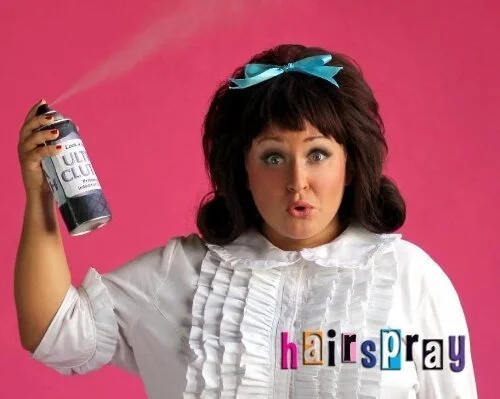 Person holding hairspray, styled like a character from Hairspray musical, with a pink background.