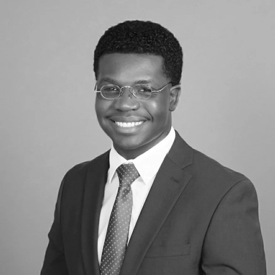 Black man wearing glasses, a suit, and a tie smiling in a studio photo.