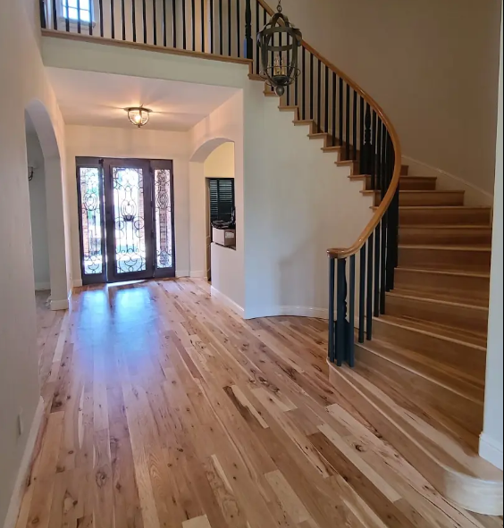 Interior view of a house entrance with light hardwood flooring, a curved staircase with black metal railing and wooden handrail, and a glass front door with decorative ironwork, visible through an open doorway.